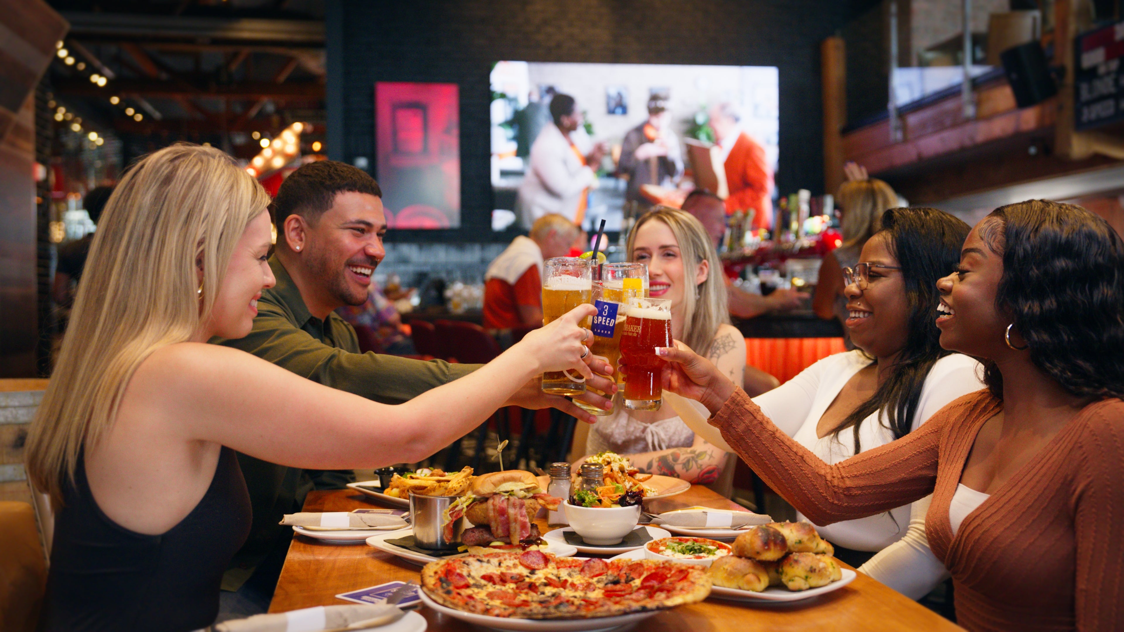 Five people sitting at a table inside Amsterdam Brewhouse, smiling and enjoying different beers and foods